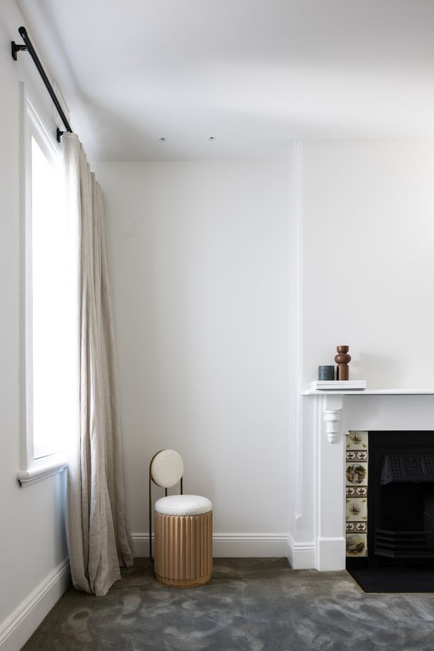 Bright, refined corner of a traditional interior featuring a tall window with full-length sheer beige curtains, white painted walls and detailed skirting and moulding, a grey carpeted floor, and a white mantel fireplace with decorative tiled surround and black firebox. A small upholstered round stool in a ribbed metallic-bronze finish sits beside the fireplace, with minimal objects arranged on the mantel.