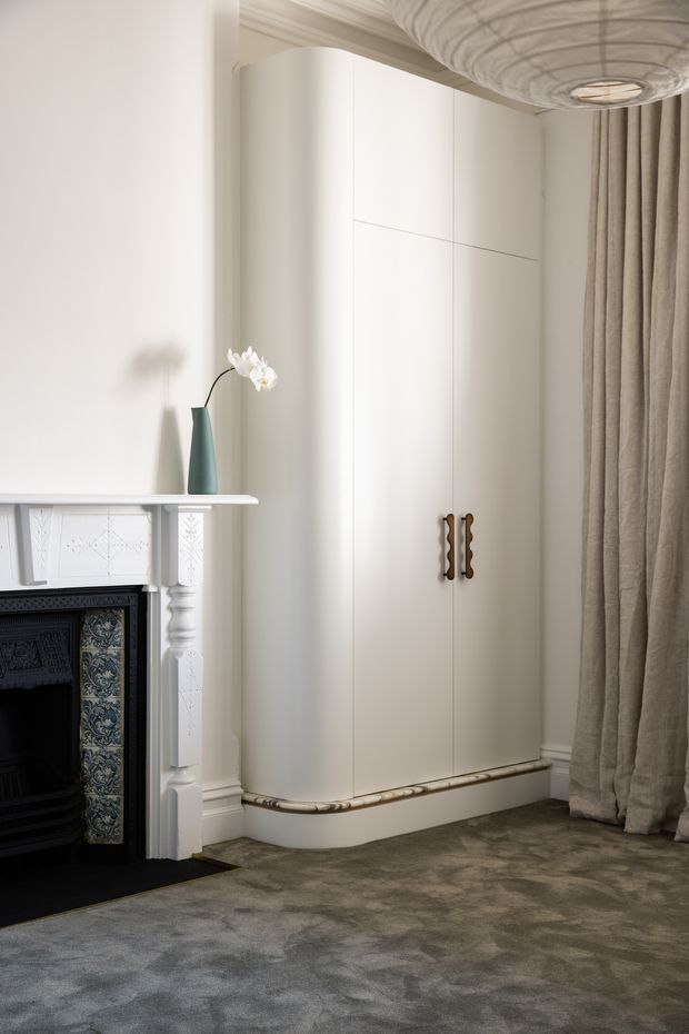 A refined traditional interior corner featuring a tall, curved built-in white wardrobe with decorative dark metal handles, beside an ornate white fireplace with patterned ceramic tile surround. Heavy taupe curtains frame the right side, while a sculptural ceiling light, plush grey-brown carpet, and a small blue-green vase add subdued detail. The space has elegant period detailing and a calm, muted atmosphere.