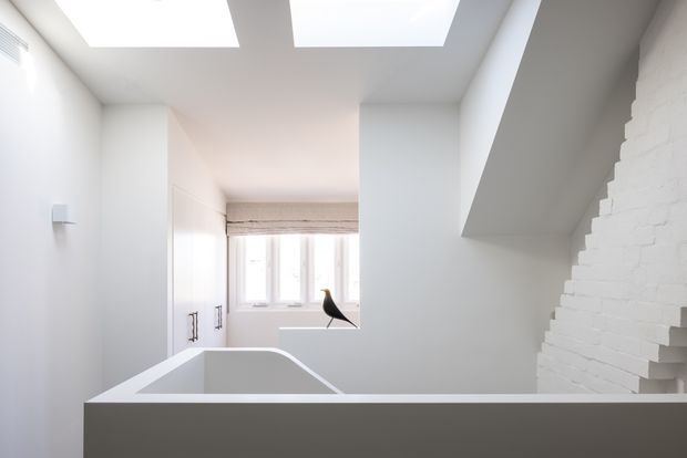 Bright minimalist bathroom viewed across a deep freestanding or built-in bathtub. The room has white walls and surfaces, a white-painted exposed brick feature wall, concealed or built-in cabinetry at left, a rear window with a light Roman blind, and two large rectangular skylights providing abundant natural light. A sculptural black bird-like object sits near the window.