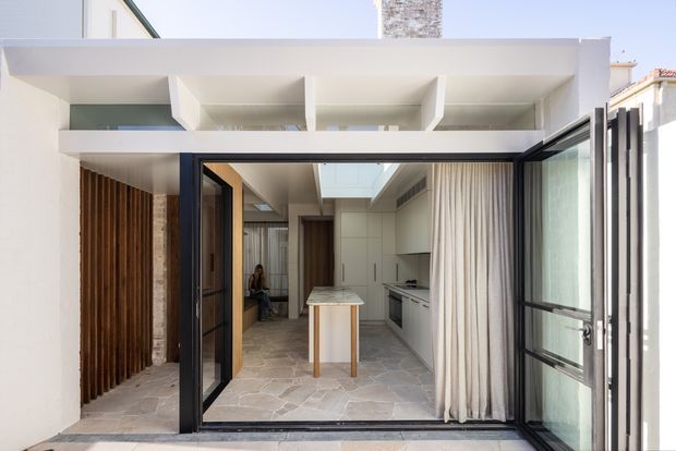 A bright contemporary kitchen extension viewed from an outdoor courtyard through wide-open black-framed glazed doors. The space features a slim central island, pale fixed cabinetry, a skylight, full-height curtains, irregular natural-stone flooring, exposed brick and vertical timber screening, with a white projecting roof structure and clerestory-like openings.