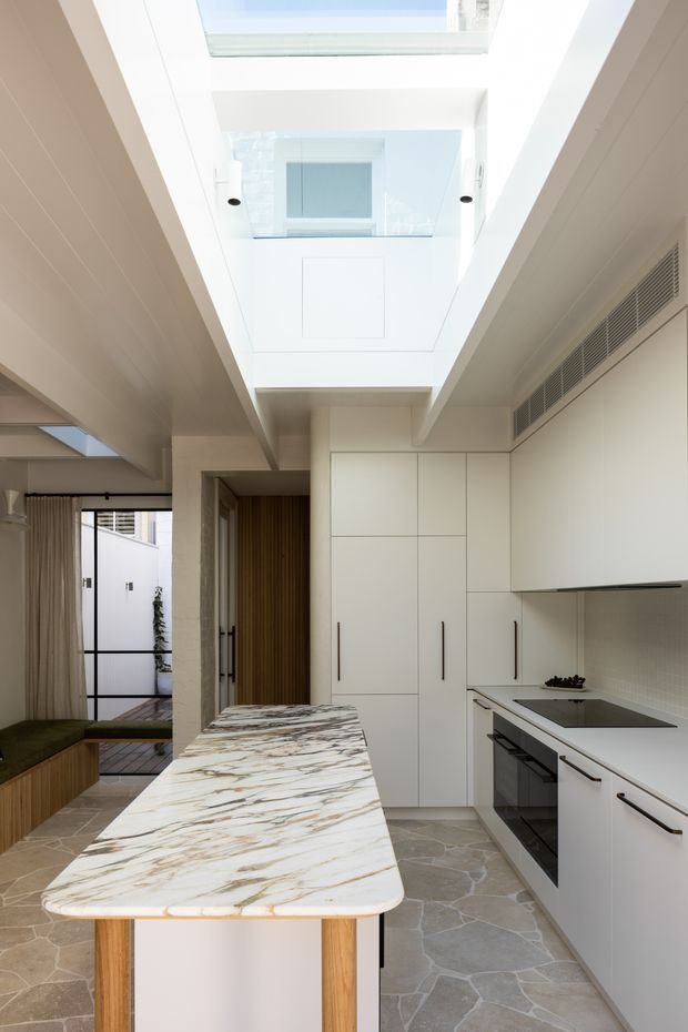 Bright contemporary kitchen viewed from the island, with a long white and marble-look island bench, streamlined white full-height cabinetry, integrated appliances, pale stone flooring, and a large overhead skylight/lightwell. A compact adjoining living or dining area is visible to the left, with timber detailing and glazing.