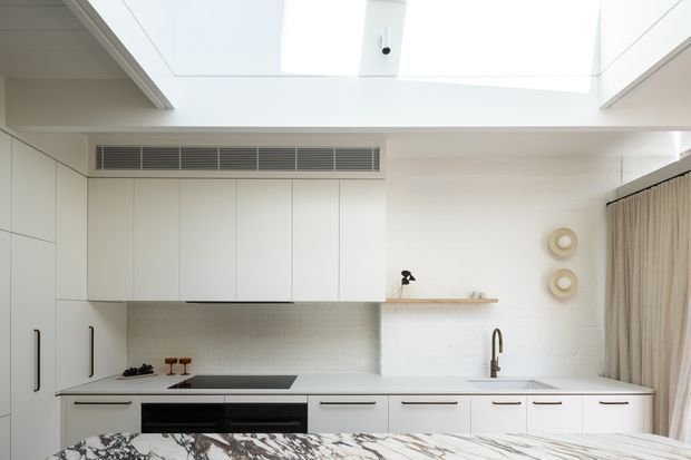 Bright minimalist white kitchen with extensive flat-panel cabinetry, integrated appliances, a long island or peninsula with a dramatic veined stone benchtop, and a skylit high ceiling. A tiled backsplash, timber shelf, slim linear air vent, and sheer curtains complete the clean contemporary interior.