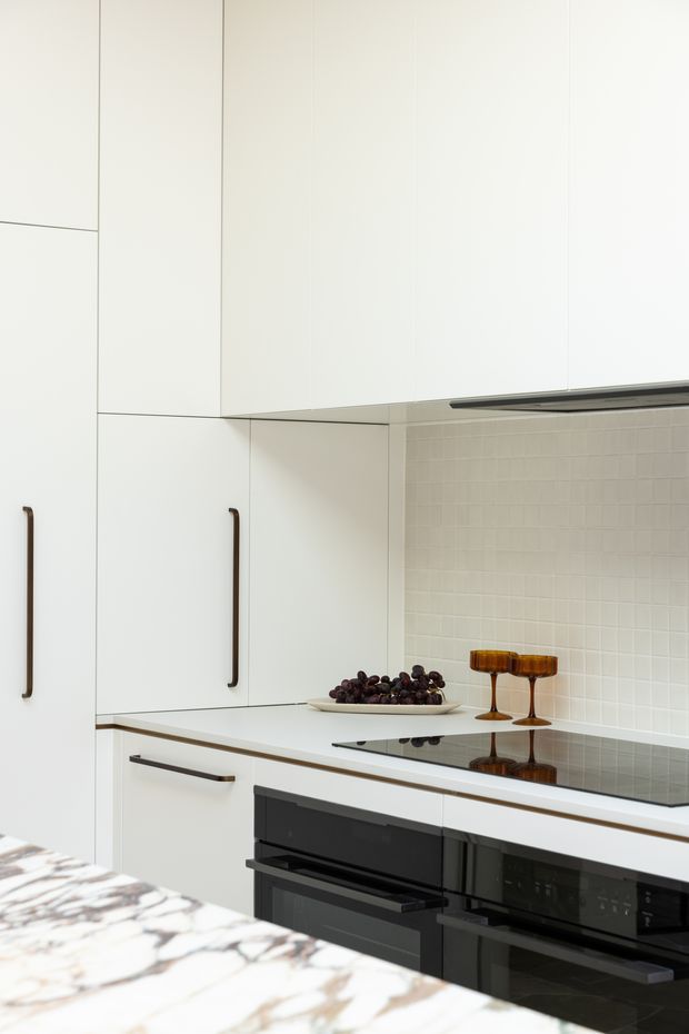 A bright, minimalist kitchen with full-height matte white cabinetry, slim dark handles, a pale tiled backsplash, white benchtops, and a black glass induction cooktop above integrated black ovens. A marble-patterned island or benchtop occupies the foreground, with small decorative glassware and a fruit bowl on the counter.