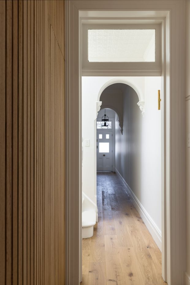 A narrow residential hallway viewed through a doorway, leading toward a glazed front door. The corridor has pale oak timber flooring, white walls and trim, an arched opening with decorative plaster detailing, and a vertical fluted timber feature wall on the left. A small portion of a white toilet is visible behind the foreground doorway, but the hallway dominates the image. Natural light enters through the front door and upper glazing, creating a bright, refined interior.