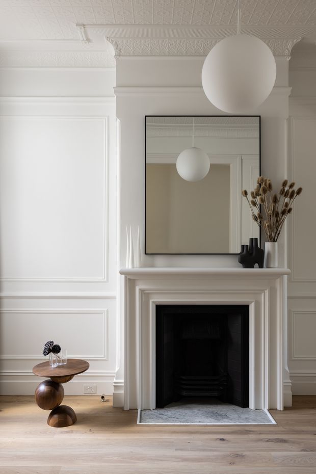 A refined living room interior centred on a traditional white-painted fireplace with layered mantel mouldings, a black firebox and pale stone hearth. The walls feature extensive decorative panel moulding, while the ceiling has ornate pressed detailing and a deep cornice. A large black-framed mirror, spherical pendant light, sculptural wooden side table and minimal decorative objects complete the calm, monochromatic space.