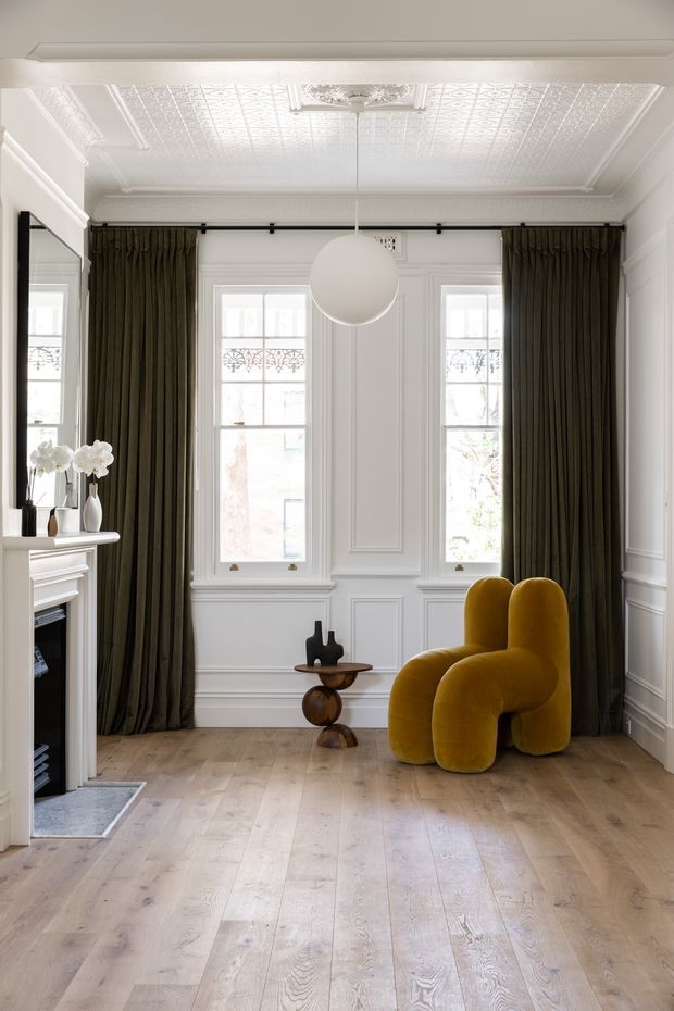 Bright, refined living room with a white-painted ornate pressed-tin ceiling, decorative wall panelling, tall sash windows, dark olive floor-to-ceiling curtains, and pale timber flooring. A sculptural mustard-yellow lounge chair provides a bold focal point, while a small sculptural side table, fireplace, and pendant light complete the elegant interior.