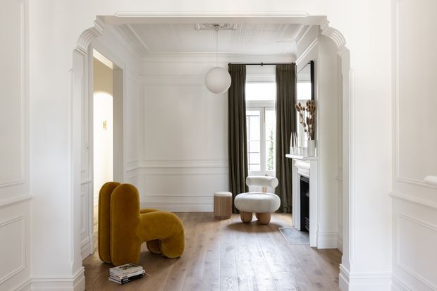 Elegant classic interior featuring a bright sitting room viewed through an ornate moulded archway. The space has white panelled walls, decorative ceiling detailing, warm timber flooring, tall dark curtains, a fireplace, and sculptural mustard and cream seating. The composition emphasizes refined architectural trim and a light-filled lounge atmosphere.