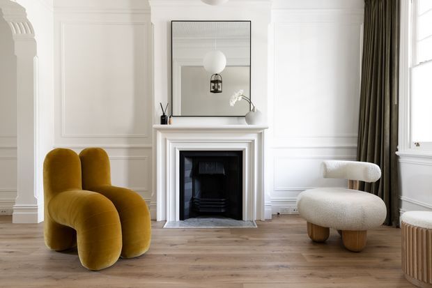 A refined living room with white decorative wall panelling, a classic fireplace surround, tall sash-style window, and warm timber flooring. Sculptural mustard velvet seating and a boucle lounge chair create a contemporary, luxurious contrast against the light traditional architectural shell. A large mirror and pendant light are positioned above the fireplace, with full-height dark curtains framing the window.