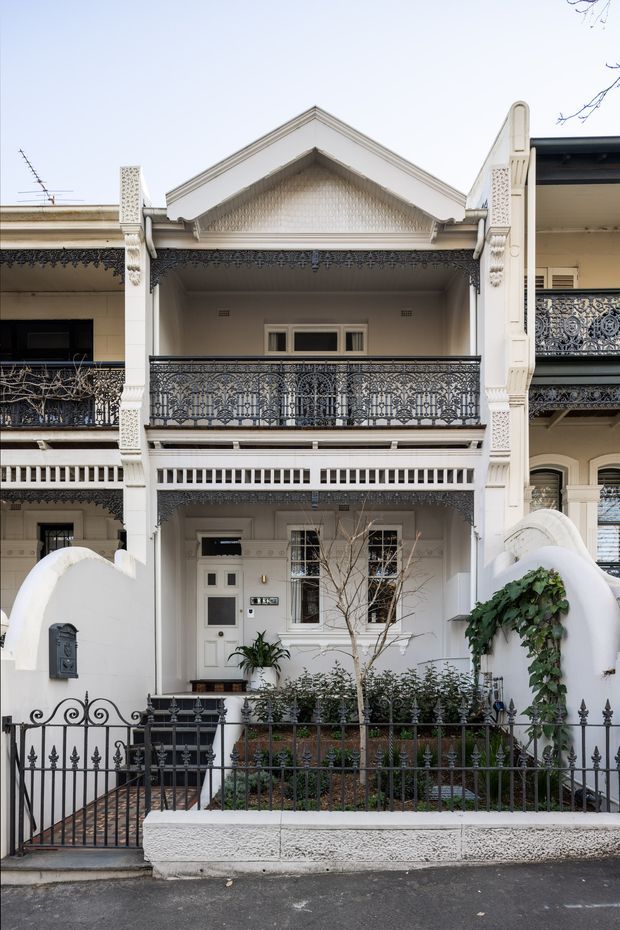 Front-on view of a narrow multi-storey heritage terrace house with an ornate white rendered facade. The building features a prominent gabled upper-level balcony, intricate cast-iron lacework balustrades and friezes, decorative columns and brackets, a recessed entry porch, and a small planted front garden enclosed by black spear-top metal fencing. Adjacent terraces are partially visible on either side.