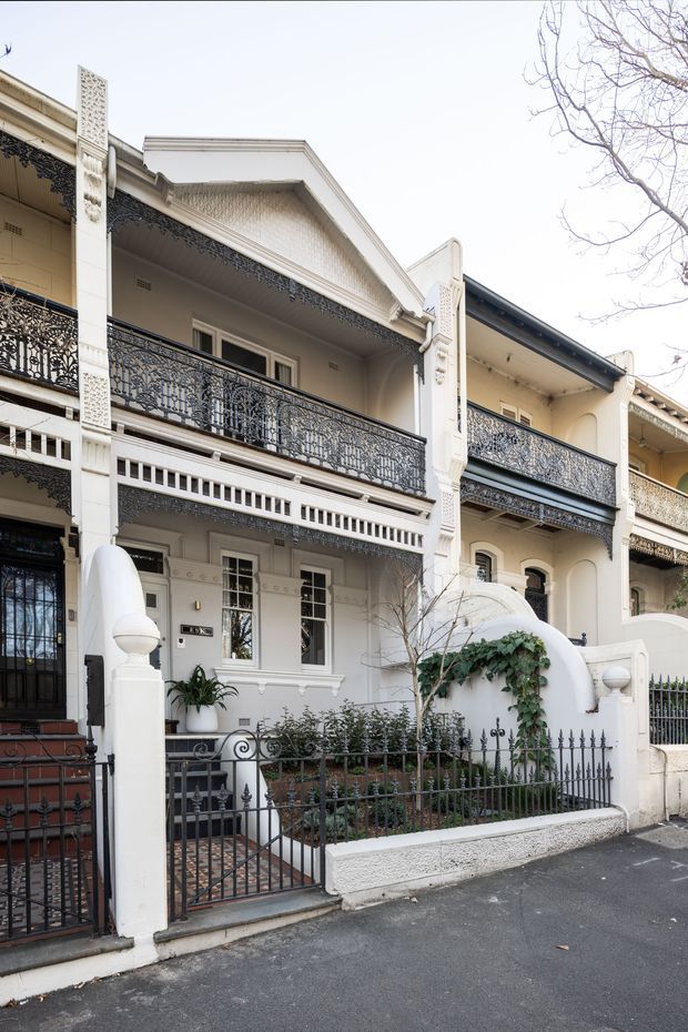 Street-facing view of an attached multi-storey terrace house with a pale rendered facade, ornate cast-iron balcony balustrades, decorative fretwork, classical columns and mouldings, arched entry detailing, and a small fenced front garden. The building has a strong Victorian-era character with later terrace-house detailing, viewed from the street.