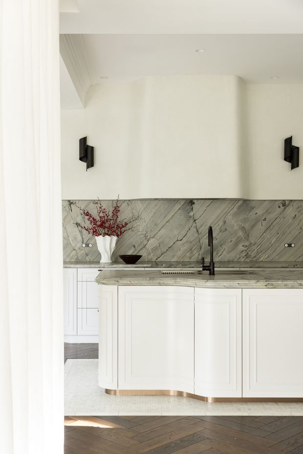 A bright, refined kitchen featuring white paneled cabinetry, a curved island with a grey veined stone benchtop and matching full-height splashback, black wall sconces and tapware, and herringbone timber flooring. A sculptural vase and branch arrangement add a restrained decorative accent.