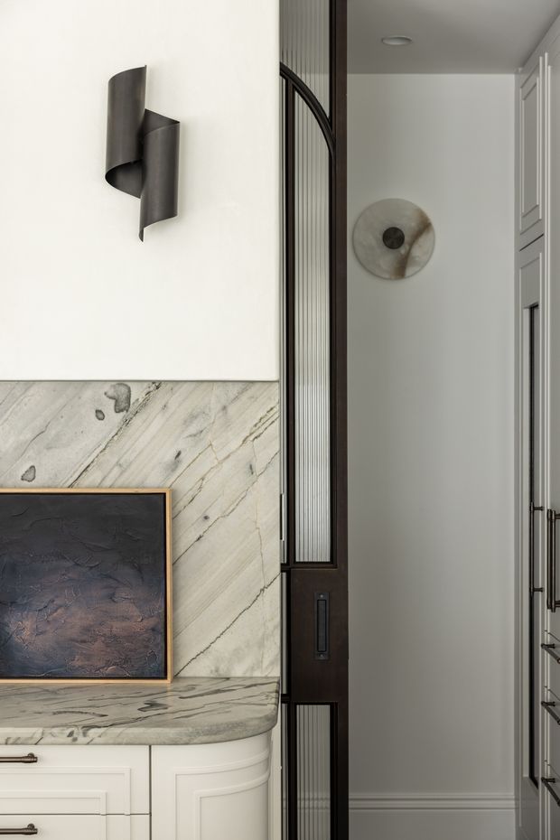 A refined interior vignette showing a marble-topped built-in console or vanity with white paneled cabinetry, a dark abstract artwork, and a sculptural black wall sconce. A dark timber-framed glazed partition with fluted or ribbed glass separates the foreground from a narrow adjacent hallway or room, where white built-in cabinetry and a circular wall artwork are visible.
