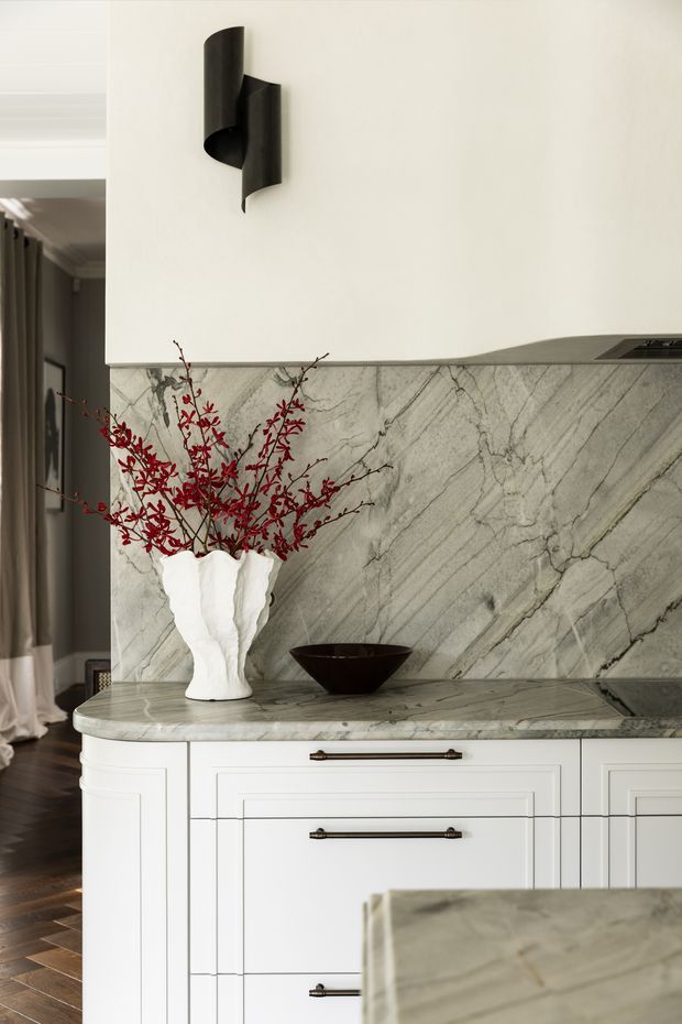 Close-up view of a refined kitchen featuring white framed cabinetry with dark metal pulls, a grey veined marble benchtop and matching full-height splashback. A sculptural black wall sconce, white organic-form vase with red branches, and dark bowl add decorative accents. Timber flooring and a curtained opening are visible at the side.