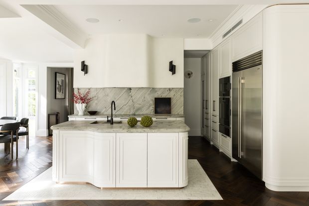 Bright, refined white kitchen with a large paneled island, marble-look splashback and benchtop, integrated cabinetry, and stainless-steel appliances. The kitchen opens to a dining area and hallway, with dark timber flooring, decorative wall lighting, and abundant natural light.