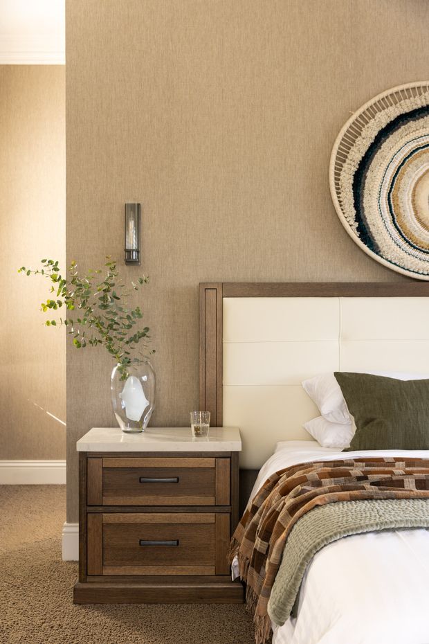 A warmly styled bedroom vignette featuring a dark timber bedside table with a light stone-look top, a cream upholstered bedhead, textured beige wall covering, wall-mounted sconce, layered neutral and brown bedding, and a circular woven wall decoration. A vase with greenery sits on the bedside table.