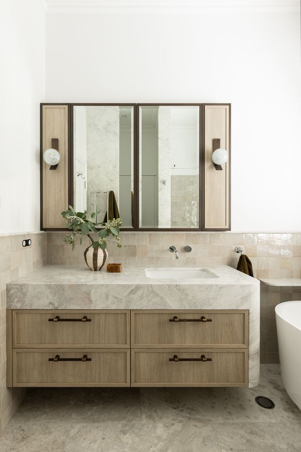 Refined contemporary bathroom featuring a double-drawer timber vanity with a pale stone countertop and integrated basin, wall-mounted brushed metal tapware, a large framed mirrored cabinet, and a freestanding white bathtub. Pale stone wall and floor finishes create a warm, calm palette, complemented by dark bronze hardware and spherical wall lights.
