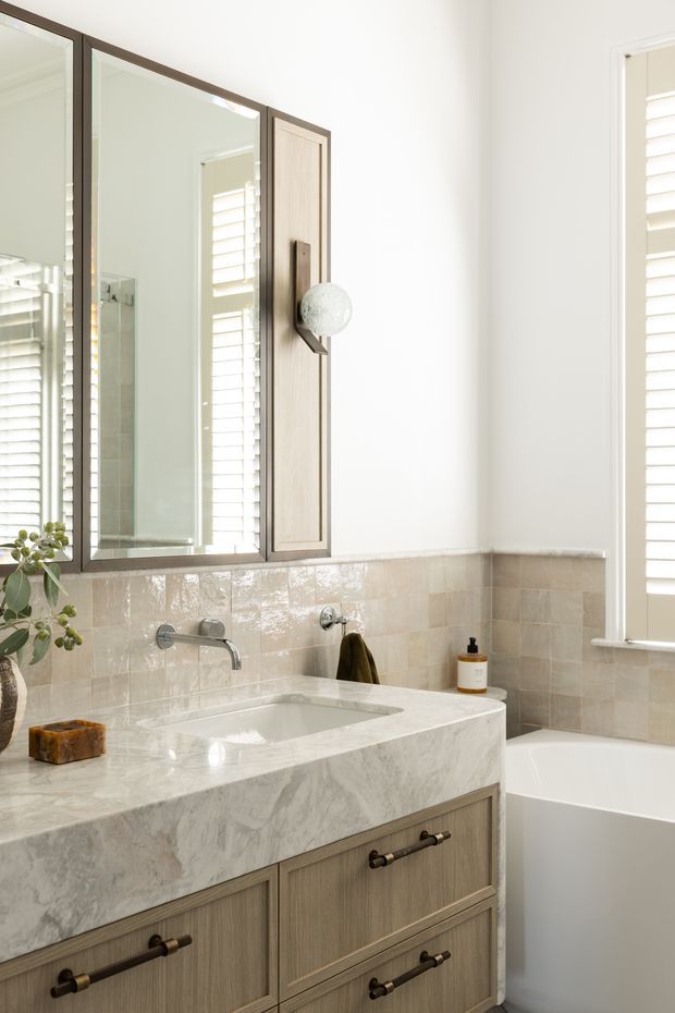 Bright, refined bathroom featuring a wall-mounted wood-look vanity with large pale marble benchtop and integrated rectangular basin, brushed metal wall-mounted tapware, glossy beige and cream tiled splashback, framed mirrored cabinetry, and a white freestanding bathtub. Plantation shutters provide filtered natural light, while dark hardware and a small plant add contrast.