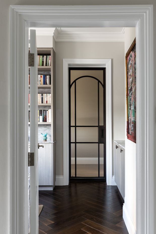A refined interior hallway framed by layered white decorative mouldings and cornices. The space features dark herringbone timber flooring, a black metal and ribbed-glass arched-panel door, built-in white cabinetry, and a partially visible bookshelf and study area beyond. A colourful framed artwork adds visual interest on the right wall.