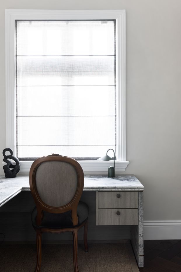 A refined home office workspace positioned beneath a large window, featuring a marble-topped built-in desk with grey wood-grain drawers, a dark wood and upholstered traditional chair, a small green desk lamp, and a sculptural black decorative object. The room has pale grey walls, white trim and window mouldings, dark herringbone timber flooring, and a woven neutral rug. Soft diffused daylight creates a calm, elegant atmosphere.