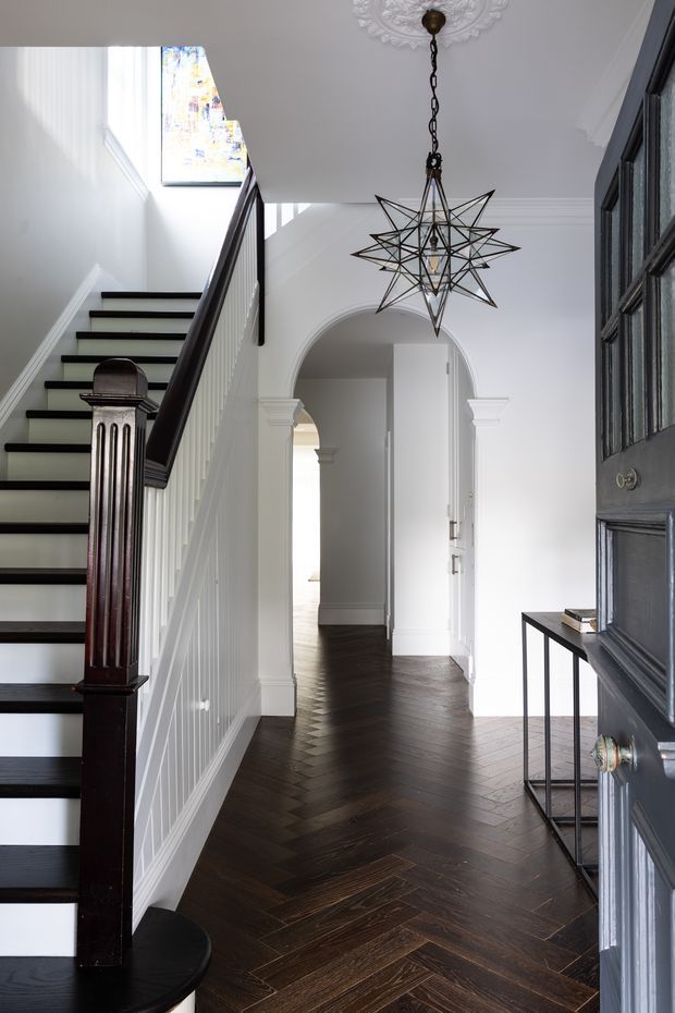 A bright, refined residential entry hall viewed from the open front door, featuring a dark-stained herringbone timber floor, white paneled staircase with dark handrail and newel post, decorative arched doorway with classical trim, white walls and ceiling, and a dramatic black geometric pendant light. A narrow console table sits beside a dark paneled entry door, while a stained-glass window brings in natural light.