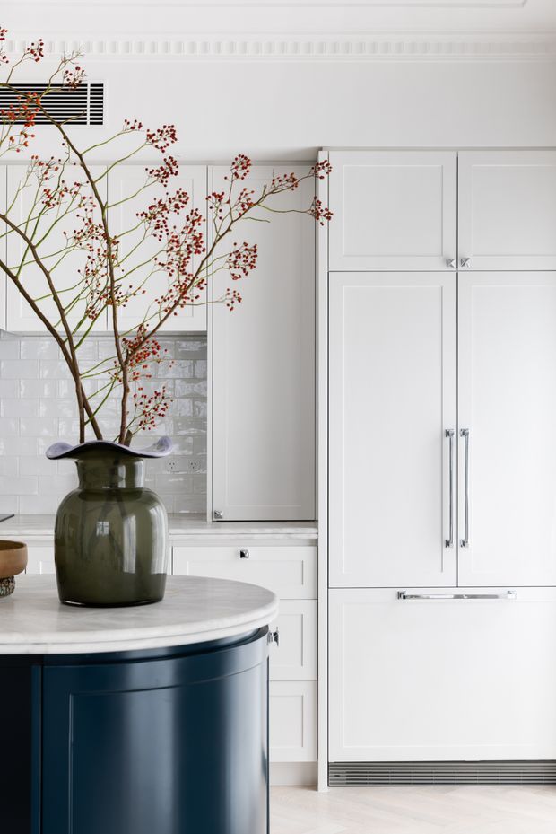 Bright, refined kitchen interior featuring a large curved dark navy island with a pale stone benchtop, white raised-panel cabinetry, glossy white subway-tile splashback, and integrated appliances. A large sculptural ceramic vase with branches forms the foreground focal point. Decorative wall moulding and a linear air vent add classic architectural detailing.