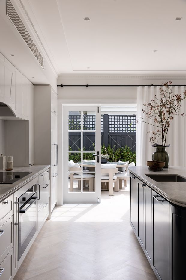 Bright, refined kitchen with white shaker-style cabinetry, dark contrasting island cabinetry, pale stone benchtops, integrated appliances, and a light herringbone timber floor. A glazed French door opens directly to a furnished outdoor dining terrace, while full-height curtains, decorative cornices, and a black curtain rail add a classic tailored character.