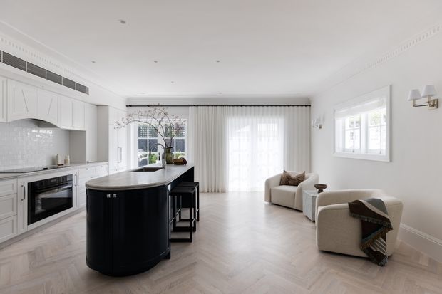 Bright, refined open-plan kitchen and living area with white Shaker-style cabinetry, a large rounded black island, pale herringbone timber flooring, sheer full-height curtains, and two cream lounge chairs. Decorative cornice, wall panelling, traditional window detailing, and brass wall sconces add a classic architectural character.