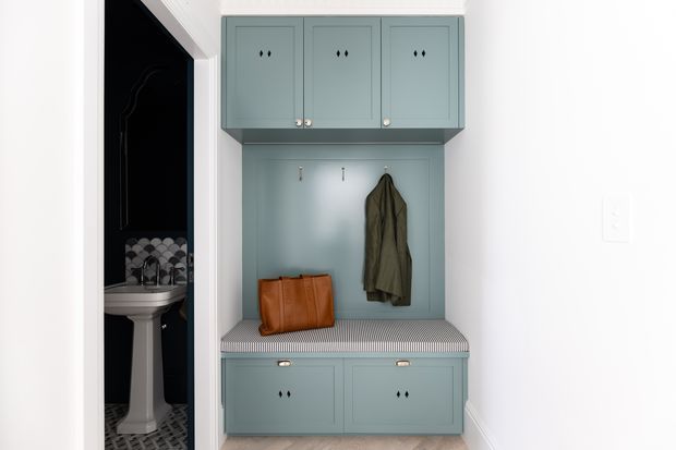 A compact built-in entryway mudroom unit with blue-grey painted cabinetry, upper storage cupboards, coat hooks, a paneled back, and a striped cushioned bench above lower drawers. A dark bathroom with a pedestal basin is visible through the adjacent doorway.