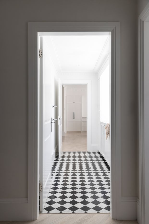 Bright, narrow residential hallway viewed through an open doorway, featuring ornate white painted wall and door mouldings, a high white ceiling with decorative cornice detailing, and a striking black-and-white checkerboard tile floor. A white built-in storage area is visible at the far end, with pale timber flooring in the foreground and a glimpse of a stair or balustrade area to the right.