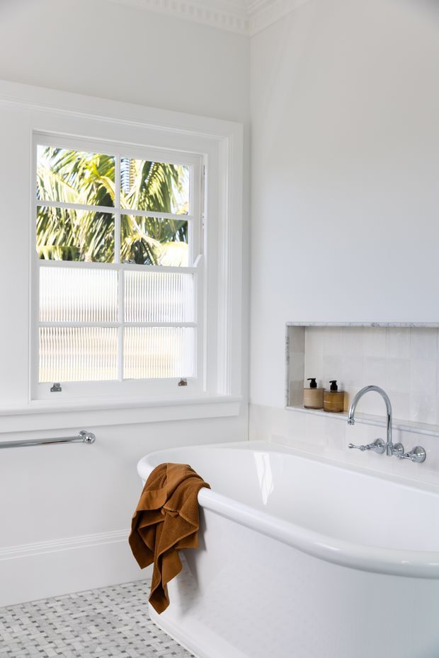 Bright, refined traditional bathroom featuring a freestanding white bathtub beneath a large sash window, a wall-mounted chrome bath tap and curved faucet, a recessed tiled niche with soap dispensers, white decorative wall mouldings, and a small-format white and grey mosaic floor. Palm foliage is visible through the textured-glass lower window panes.