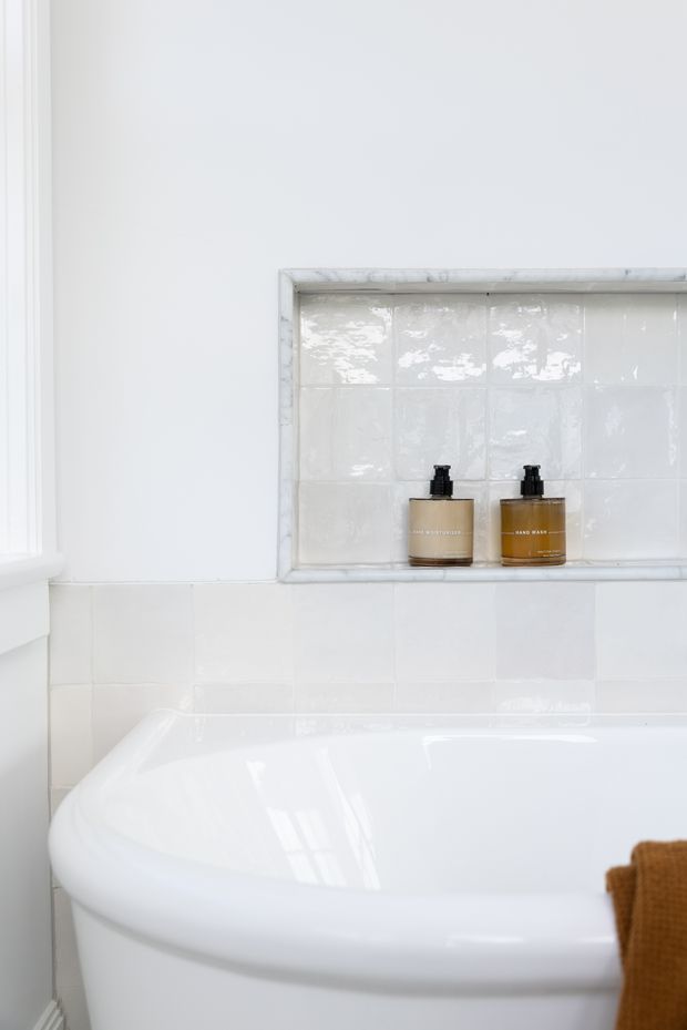 Close-up of a bright, predominantly white bathroom featuring a glossy white bathtub in the foreground, white tiled walls, and a recessed tiled shelf holding two amber soap dispensers. The space has a clean, simple, understated appearance with white-painted trim and polished ceramic surfaces.