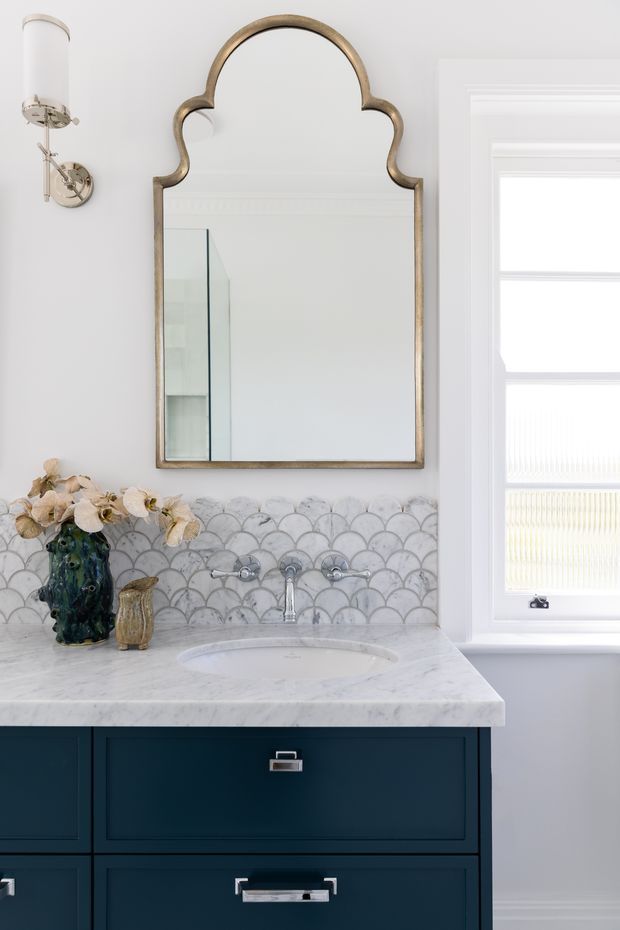 A refined bathroom vanity with deep navy-blue cabinetry, a white marble countertop and integrated oval sink, chrome tapware, and a scalloped grey-and-white tile splashback. An ornate aged-metal framed mirror hangs above the vanity beside a traditional wall sconce and a white-framed window with blinds. Decorative vases and flowers add a small ornamental accent.
