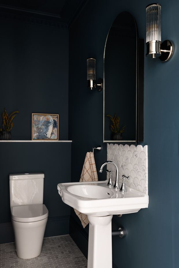 A compact, dark blue bathroom featuring a white pedestal basin with polished chrome tapware, a white toilet, scalloped white wall tiles behind the basin, a tall arched mirror, and elegant ribbed wall sconces. A small shelf with artwork and a plant adds decorative detail. The floor is finished in small grey mosaic tiles, creating a moody yet refined interior.