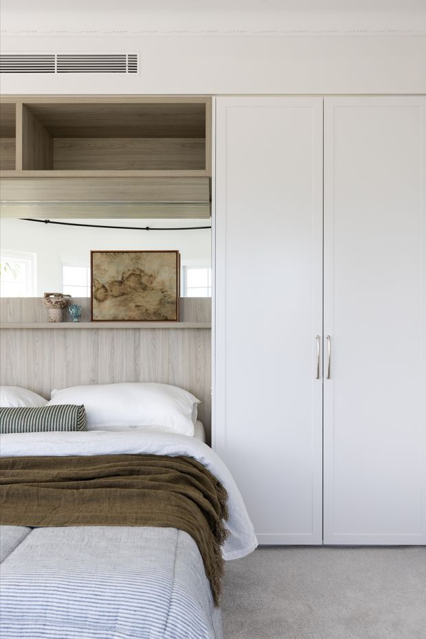 A calm, light-filled bedroom featuring a built-in bed integrated into pale timber joinery, a large white built-in wardrobe with slim metal handles, mirrored shelving, layered white bedding, and a muted olive-brown throw. The room has soft carpet, restrained neutral colours, and simple refined detailing.