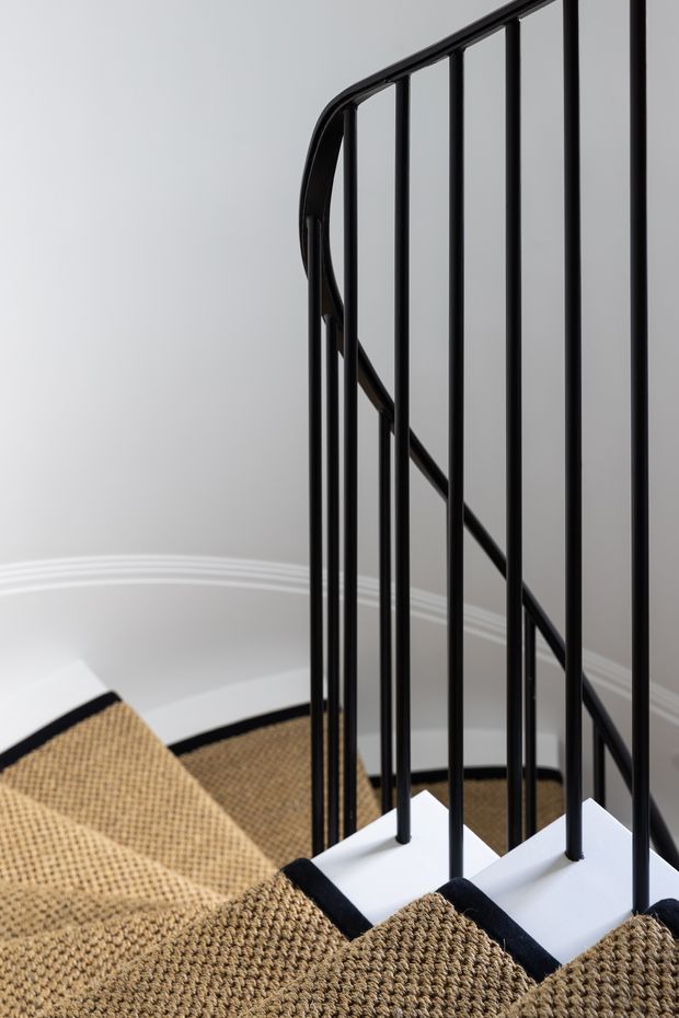 Close-up architectural photograph of a staircase with white treads, thick woven beige carpet runners, and a slim black metal balustrade featuring vertical rods and a curved handrail. The stairwell has light grey walls and white decorative skirting, creating a clean, refined contemporary interior.