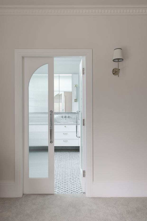 A refined traditional hallway or landing looking through a partially open, white-trimmed glazed door into a bright bathroom. The hallway has taupe painted walls, ornate decorative cornice and dado moulding, a wall-mounted traditional sconce, and soft grey carpet. The bathroom beyond features a white vanity, marble-look countertop, mirrored cabinetry, a chrome tap, and patterned geometric floor tiles. The door incorporates vertically ribbed translucent glass and elegant panel detailing.