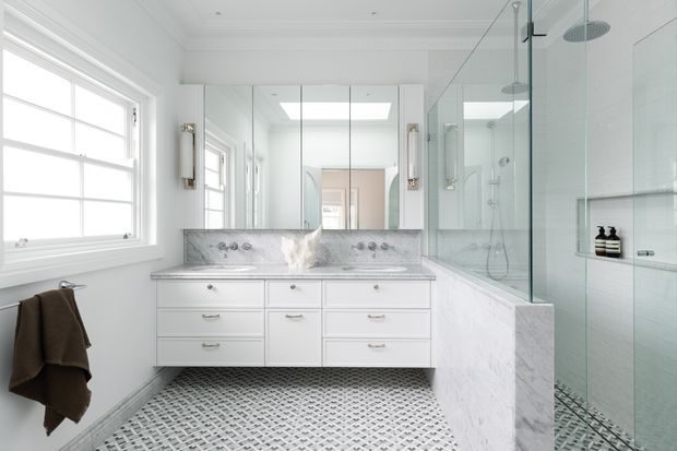 Bright, refined bathroom featuring a double vanity with white painted cabinetry, marble benchtop and integrated backsplash, mirrored wall cabinets, a freestanding-style shower enclosure with clear glass, and a small-format patterned tile floor. A large window and skylight provide abundant natural light, while brass-toned wall sconces add warmth.