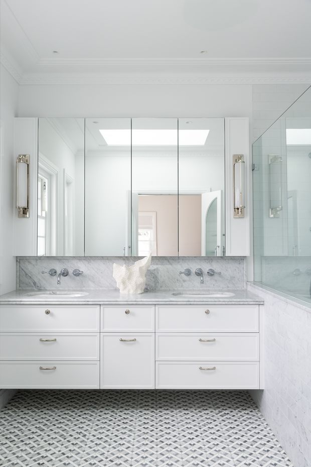 Bright, refined bathroom featuring a wide double vanity with white shaker-style cabinetry, marble benchtop and backsplash, mirrored cabinet doors, twin undermount basins, polished metal wall-mounted tapware, decorative wall sconces, white tiled walls, and a geometric grey-and-white patterned floor. A glazed shower enclosure is visible at right, with a skylight and detailed ceiling cornice adding natural light and architectural character.