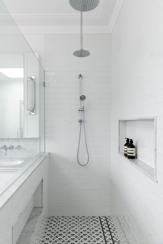 Bright, predominantly white bathroom with a walk-in shower, white subway-tile walls, patterned geometric floor tiles, a glass shower partition, a marble-look vanity with integrated basin, recessed wall niche, and ornate decorative ceiling detailing. Chrome shower fixtures and a large rain shower add a refined traditional feel.