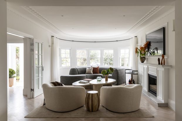 Bright, refined living room in a period-style home, featuring a curved bay window with sheer curtains, ornate ceiling cornice, white fireplace surround, pale timber flooring, a grey sofa, rounded cream lounge chairs, and a round coffee table. French doors open toward an adjacent space, while a large mirror and restrained decorative styling add a polished, contemporary-classic feel.