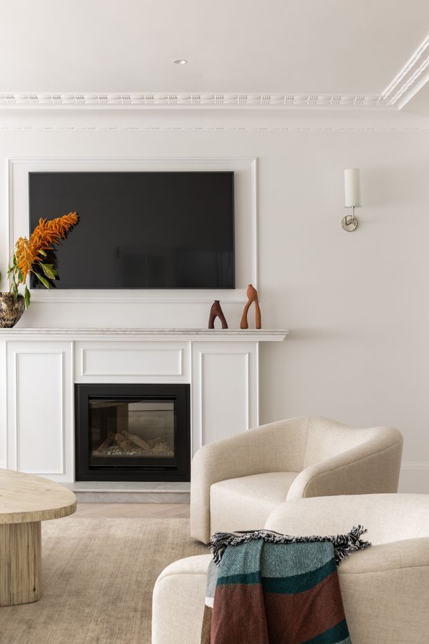 A refined living room with a white decorative fireplace surround and mantel, wall-mounted television, ornate ceiling cornice, sculptural wall sconce, neutral upholstered armchairs, a light timber side table, and a muted striped throw. The room has a calm, warm, elegant traditional-contemporary character.