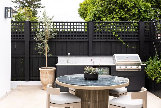 A refined outdoor dining and entertaining area with a round stone-topped dining table and upholstered chairs, set beside a built-in outdoor kitchen with barbecue, sink and dark cabinetry. The space is enclosed by a black timber privacy fence with lattice screening, complemented by potted planting and a wall-mounted exterior light.