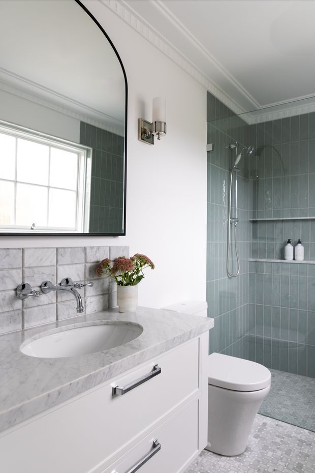 A bright, refined bathroom with a white marble vanity and integrated oval basin, chrome wall-mounted tapware, white cabinetry, a large arched-edge mirror, and traditional decorative cornices. The shower enclosure is finished in vertically oriented muted green tiles with a built-in niche and chrome shower fittings. A white toilet and small mosaic floor tiles complete the space.