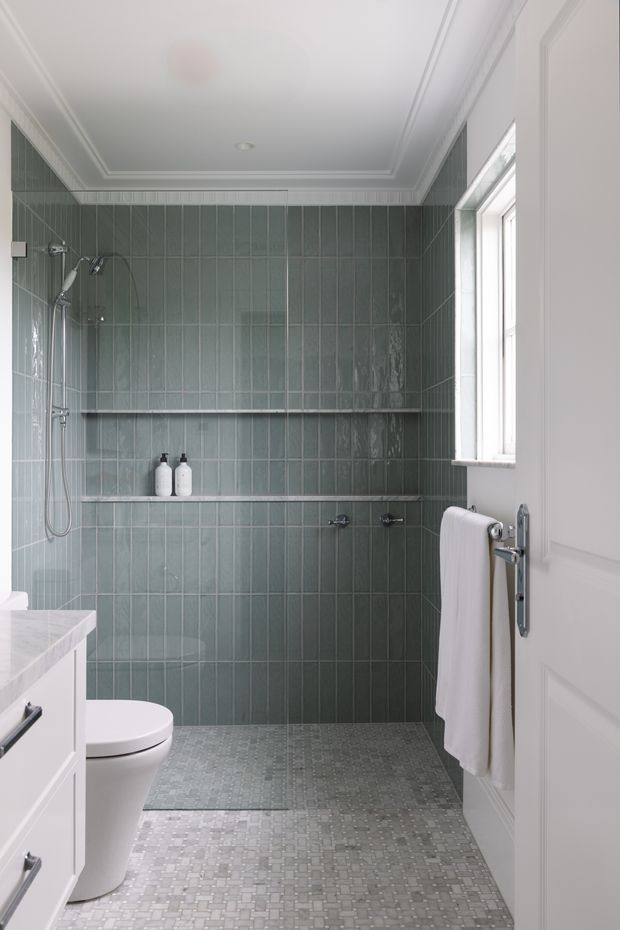 A compact, elegant bathroom with a walk-in shower, toilet, white vanity, towel rail, and a large window. The walls are clad in vertically arranged glossy muted green-grey tiles, while the floor features small mosaic tiles in cool grey tones. White moulded door and ceiling trim add a traditional architectural detail, complemented by simple chrome fixtures and recessed lighting.