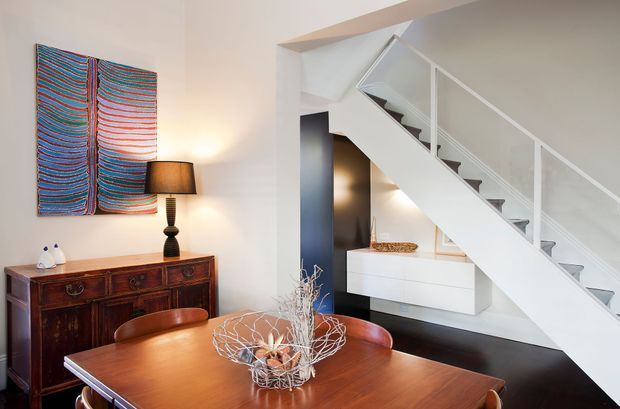 A refined open-plan dining area with a polished dark timber dining table and matching chairs, a vintage-style timber sideboard, colourful abstract wall art, and a table lamp. A striking white stair with glass balustrade rises beside built-in white cabinetry, creating a strong contemporary architectural feature. The palette combines warm timber and dark flooring with crisp white walls and joinery.
