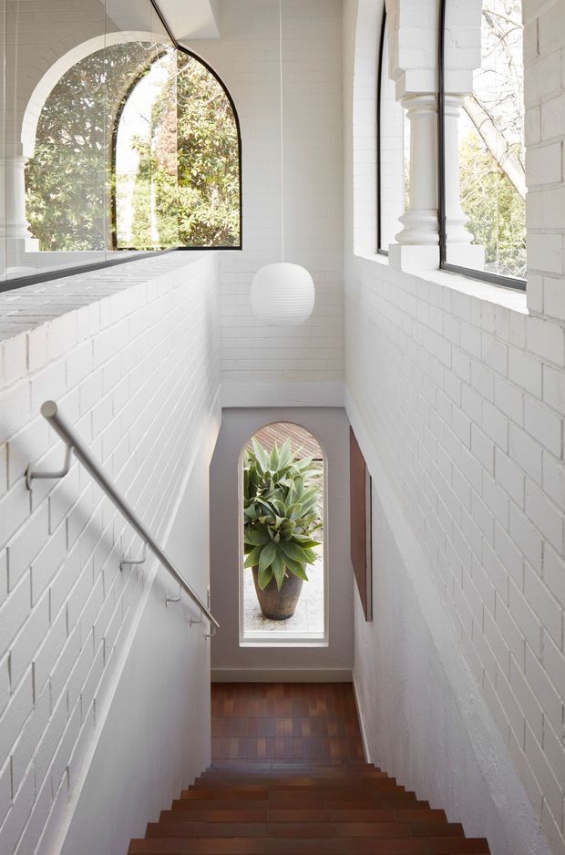 A bright, narrow stairway descending through a white-painted brick interior. The stairwell features warm timber stair treads and flooring, slim metal handrails, arched windows and a tall arched glazed opening overlooking a planted courtyard. Decorative white column detailing and a spherical pendant light add character, while abundant natural light creates an airy vertical space.