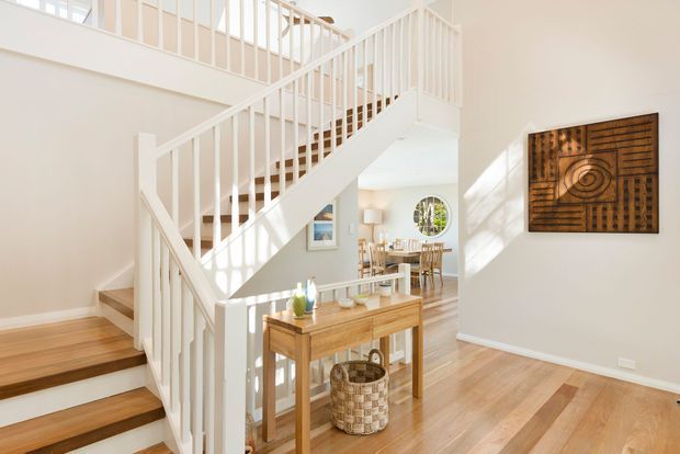 Bright, airy interior view focused on a timber staircase with white balustrades and railings, opening onto a landing and dining area. Light timber flooring, white walls, a narrow timber console table, woven basket storage, and a framed carved timber artwork create a warm, clean residential setting.