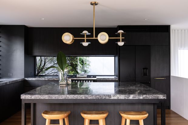 A sophisticated contemporary kitchen viewed head-on, featuring a large dark stone island with three timber stools, black fixed cabinetry, a matching dark stone benchtop and splashback, and a wide horizontal window above the cooktop. A sculptural brass-and-opal-glass pendant light hangs centrally over the island. The palette is dark and dramatic, balanced by warm timber seating and greenery visible through the window.