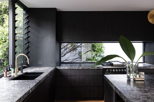 Contemporary dark-toned kitchen with an L-shaped layout and central island, featuring black timber-look cabinetry, polished grey natural-stone benchtops, a stainless-steel range cooker, and a deep undermount sink with a curved brass tap. Full-height windows and horizontal louvres bring in abundant greenery and daylight, while a sculptural wall light and indoor plant add restrained decorative accents.