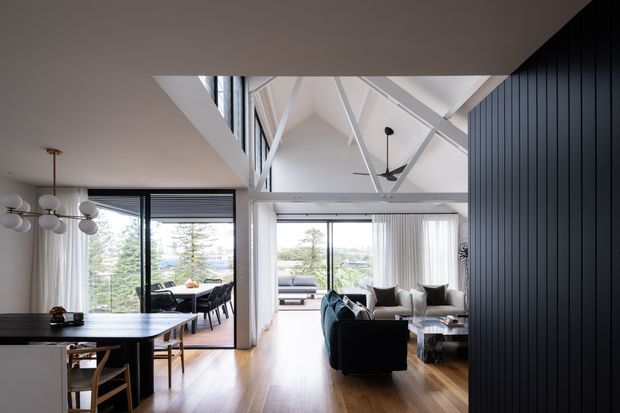 A bright, open-plan contemporary interior combining a dining area, living room, and visible upper-level void. The space features warm timber flooring, black vertical wall panelling, expansive glazed openings, white structural beams and angled ceiling forms, and a sculptural multi-globe pendant above the dining table. A living area is visible beyond the central opening, with landscaped views through the glazing.